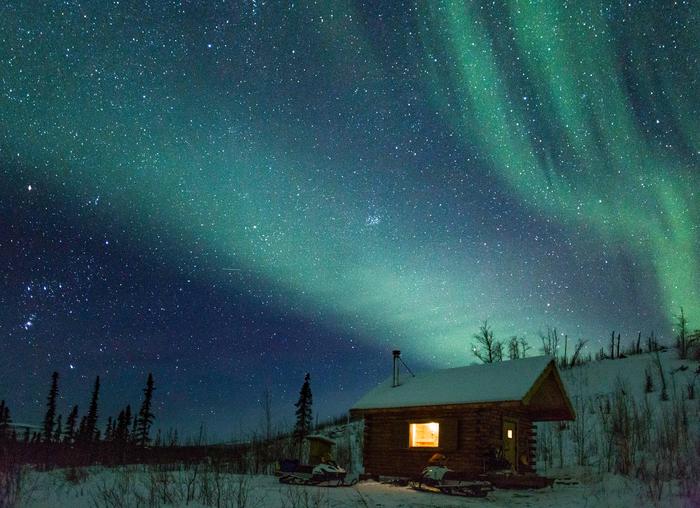 A cabin next to a low hill beneath northern lightsNorthern lights at Wolf Run Cabin.