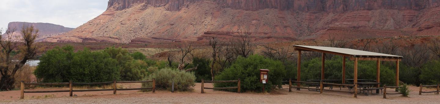 The Lower Onion Creek Group Site A has a shade shelter with picnic tables underneath and fence separating tent camping area from parking area. Tall, red rock cliffs in the distance. 