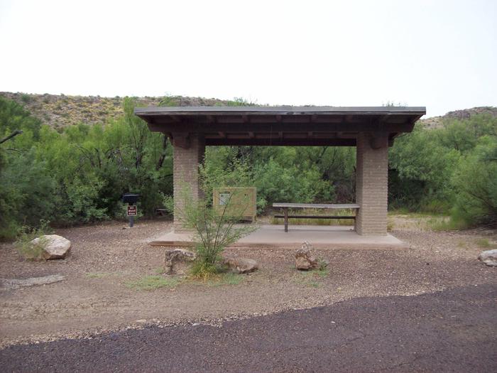 A view of the open shade shelter at Site 13 is shown with a picnic table attached underneath the shelter. There is a bear box just behind the platform that the shelter is attached to. View of the shade shelter at Site 13