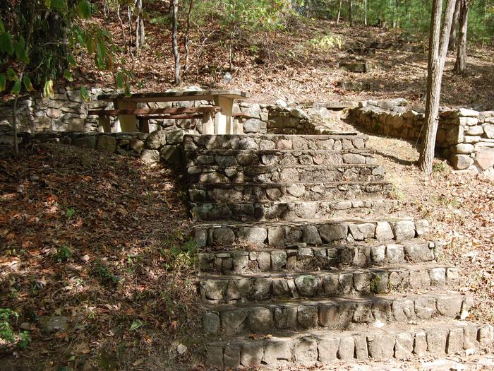 12 Stairs Site is in a wooded area of the campground. Stairs lead up to a shaded picnic area and tent pad. 