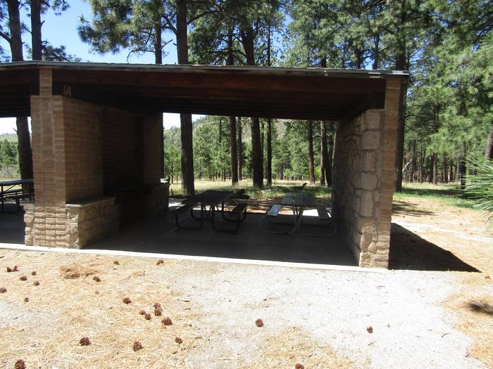 Covered rock shelter with picnic table and grills inside surrounded by pine treesCampsite A in Ponderosa Group Campground includes access to a covered cooking shelter with grills and picnic table.