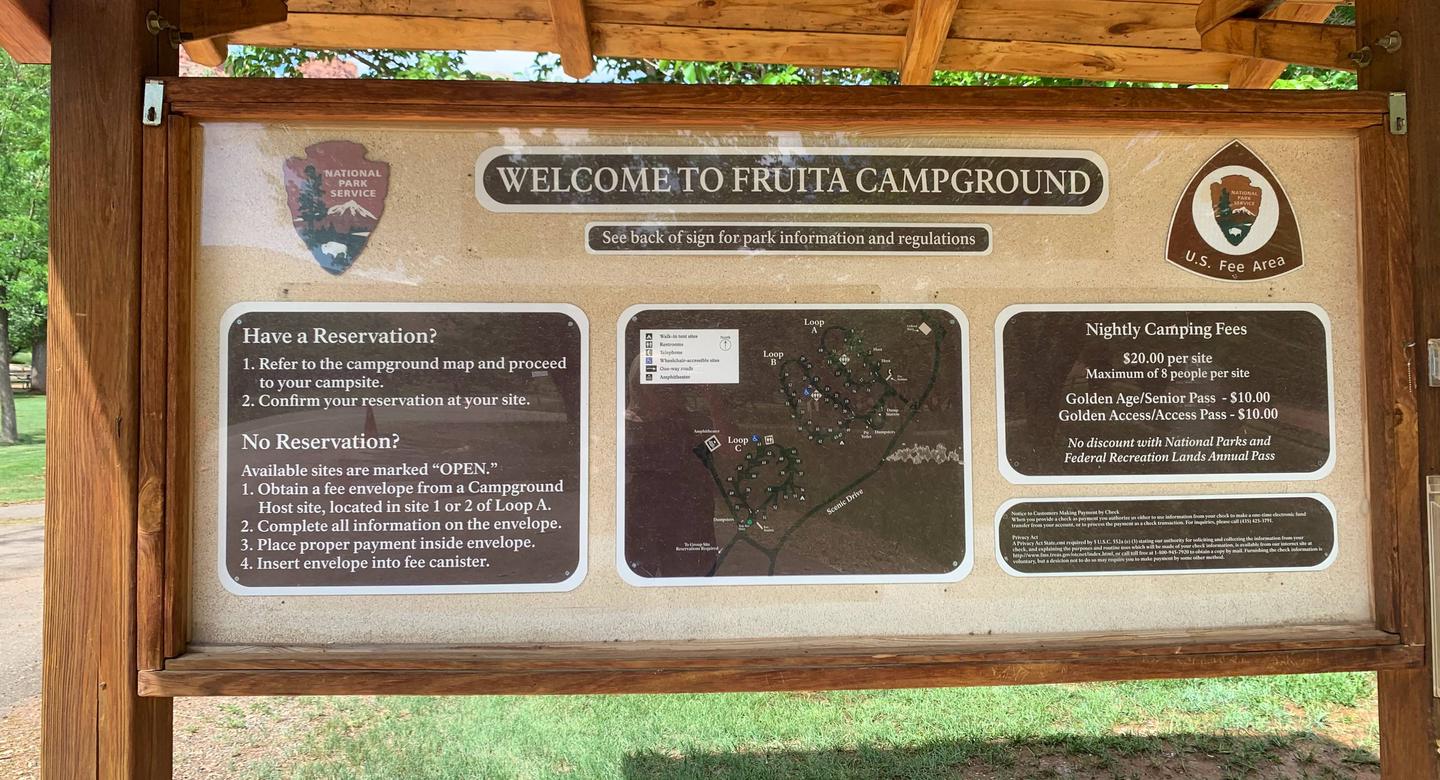 A Bulletin Board with 8 signs in it. Going clockwise starting at the top of the clock, the signs are as follows: "Welcome to Fruit Campground" ; "See back of sign for park information and regulations"; The NPS Arrowhead, underneath is "U.S. Fee Area"; "Night Camping Fees. $20.00 per site; Maximum of 8 people per site. Golden Age/Senior Pass- $10.00; Golden Access/Access Pass- $10.00. No discount with National Parks and Federal Recreation Lands Annual Pass; Privacy Act sign; Map of the campground; "Have a Reservation? 1. Refer to the campground and map and proceed to your campsite. 2. Confirm your reservation at your site. No Reservation? Available sites are marked "OPEN." 1. Obtain a fee envelope from a Campground Host site, located in site 1 or 2 of Loop A. 2. Complete all information on the envelope. 3. Place proper payment inside envelope. 4. Insert envelope into fee canister; NPS Arrowhead/Loop C Campground Kiosk