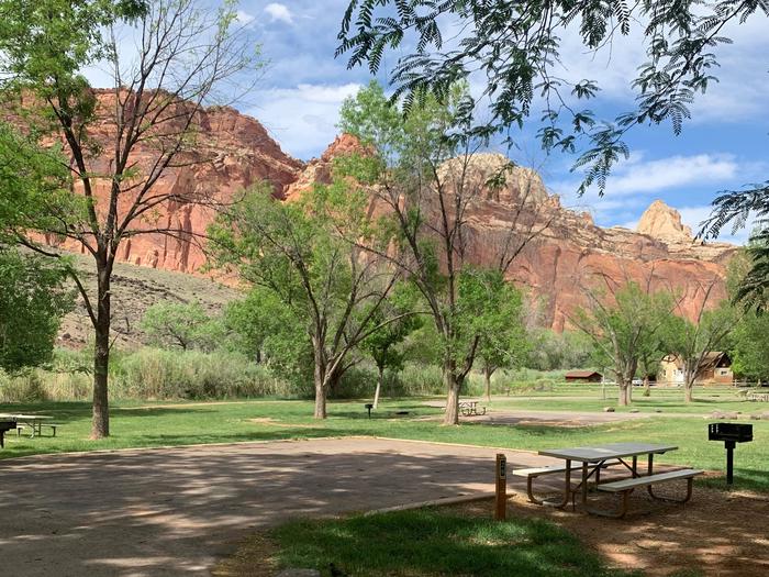 A paved driveway. Facing the end of the driveway, a picnic table and grill are to the right side. There are a few trees in the backgrounds. Tall red cliffs are in the very back across the image.Site 28, Loop B in summer.
Paved Dimensions: 30' x 30'
