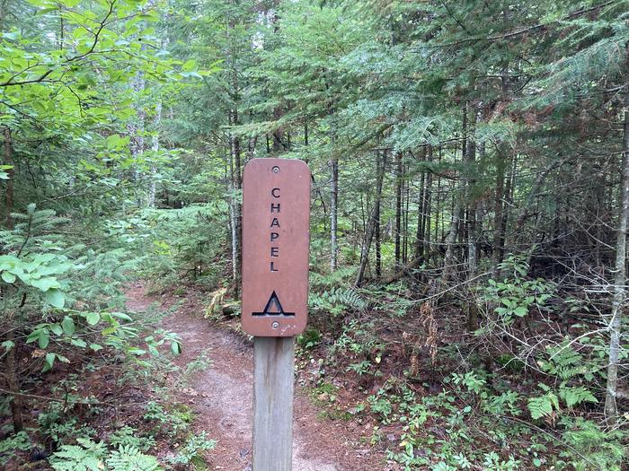 A brown sign on a pole on a wooded trail saying Chapel with a campground symbol.