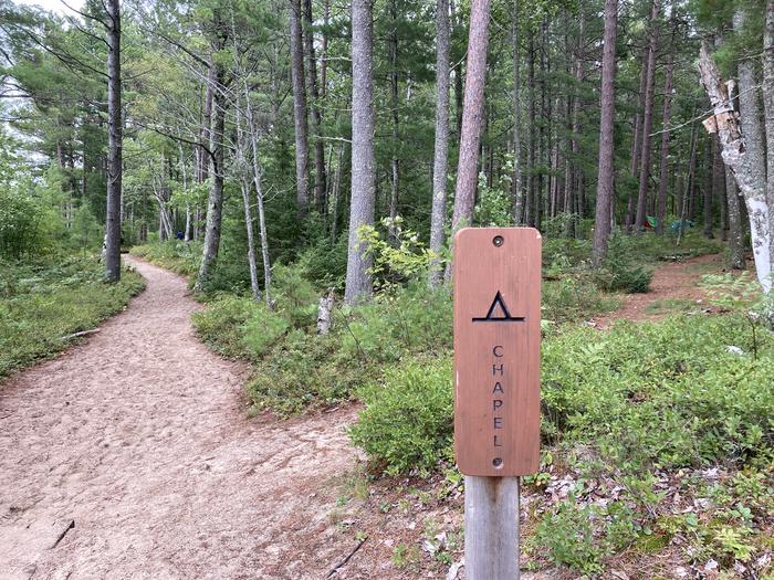 A brown post on a wooded trail saying Chapel with a campground symbol on it.