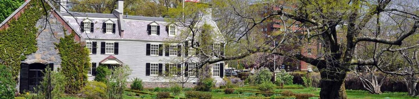 A side view of two buildings. On the left, there is a small stone building covered with wisteria and next to it a large home with gray siding, white trim, and black shutters. In the foreground is a garden with boxwoods and a large yellowwood tree on the right. A view of Old House at Peace field from the formal garden.