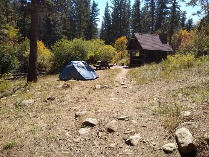 A sheltered site with a tent, picnic table, and fire place with fall foliage and pines in the background.Panchuela Site 1