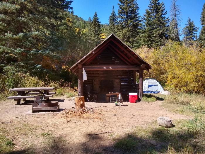 A shelter unit with a fireplace, ppicnic table, and grill along with a tent, cooler and fall foliage.Panchuela Site 5