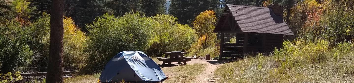 A sheltered site with a tent, picnic table, and fire place with fall foliage and pines in the background.Panchuela Site 1 Hero Image