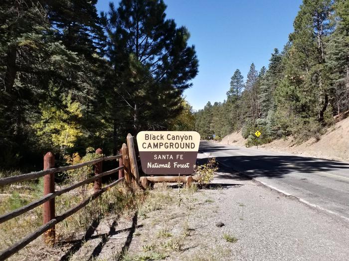 A campground sign with the words Black Canyon with a road and trees in the background.Campground sign