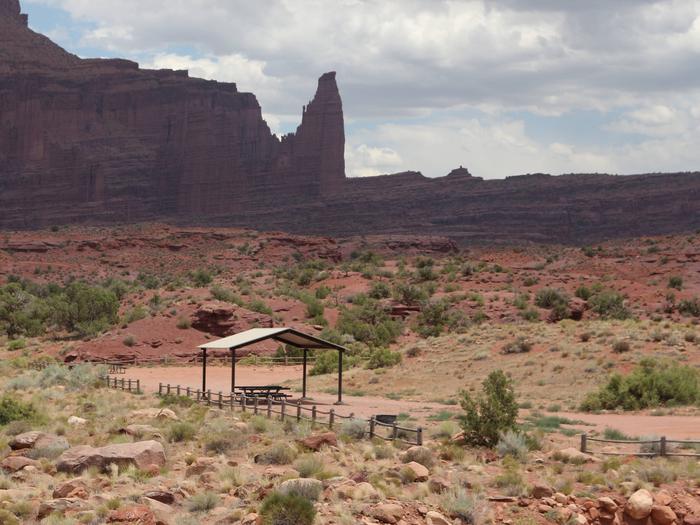 The Upper Onion Creek Group Site B has a large shade shelter with picnic tables underneath, surrounded by undulating, shrub-covered desert land. In the distance, the Fisher Towers area can be seen.