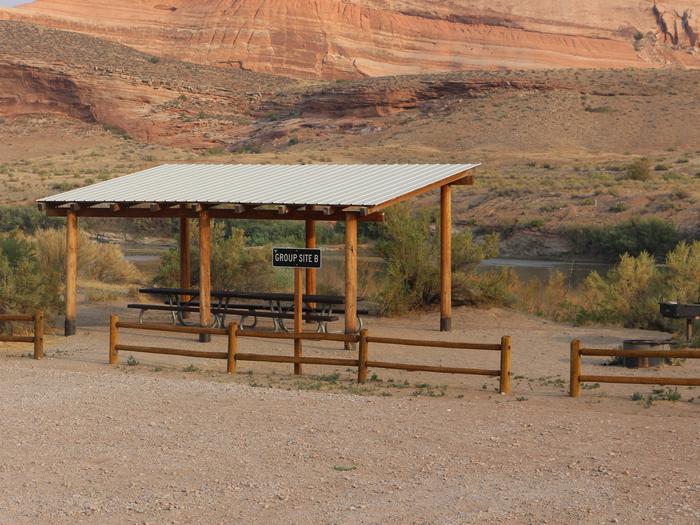 Close up of the Dewey Bridge Group Site B shade shelter, picnic tables, and parking area. The Colorado River runs adjacent to the site with tall, red rock cliffs in the distance. 
