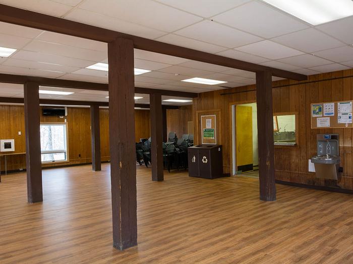 Interior view of dining hall shows 5 large wooden beams, oak laminate flooring, stacked chairs, 6 ft. tables, bottle filling station, waste/recycle bin, and doorway to dishwashing room.