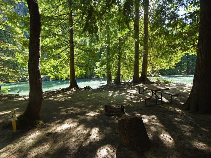 Picnic table and fire grill with trees and river in the background. Campsite near Stehekin River. 