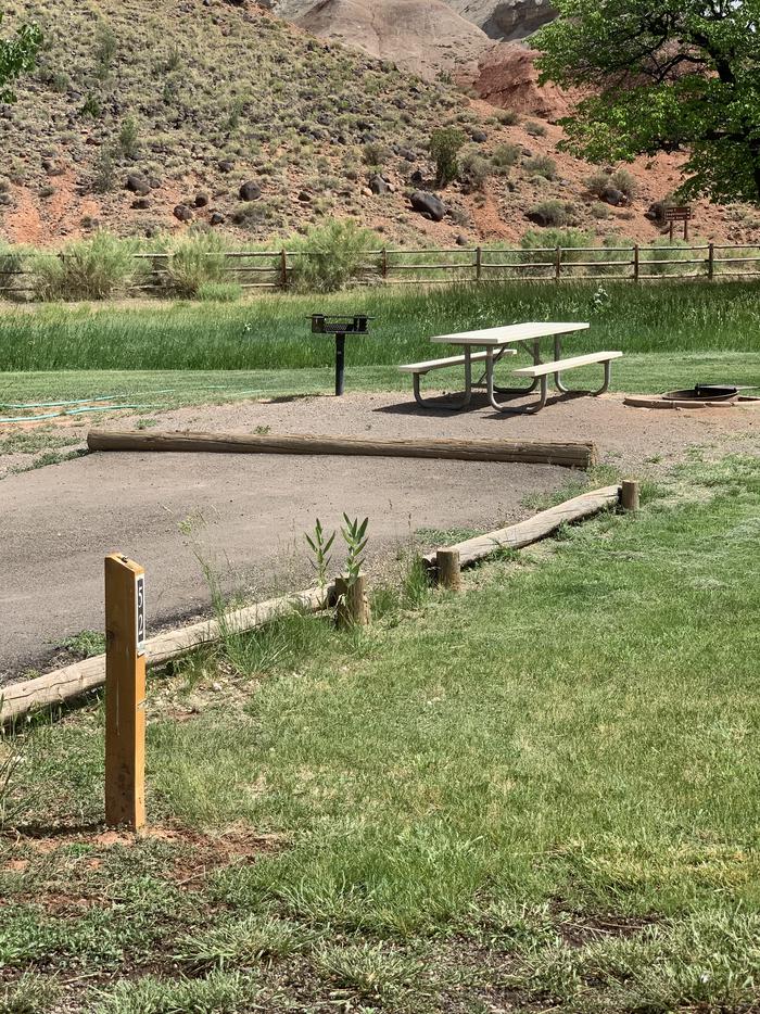 A paved driveway. Facing the end of the driveway, a picnic table, fire pit, and grill are directly behind the site. A fence is the background going across the image. Red-colored cliffs rise above the fence.Site 52, Loop C in summer.
Paved Dimensions: 18' x 42'.