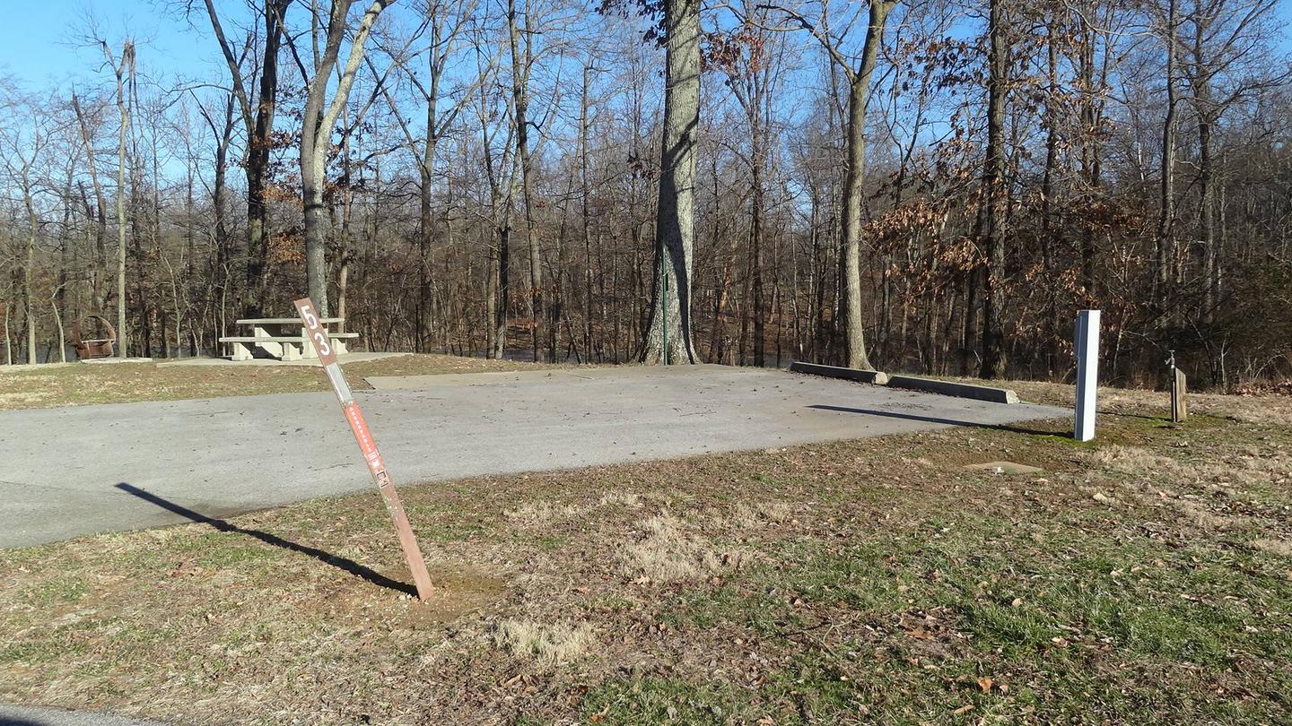 his site is nestled in the trees providing shade. The picnic table and fire pit are to the left of the concrete pad. The picnic table sits on a paved pad. This is a full hookup site and all hookups are located on the right. 