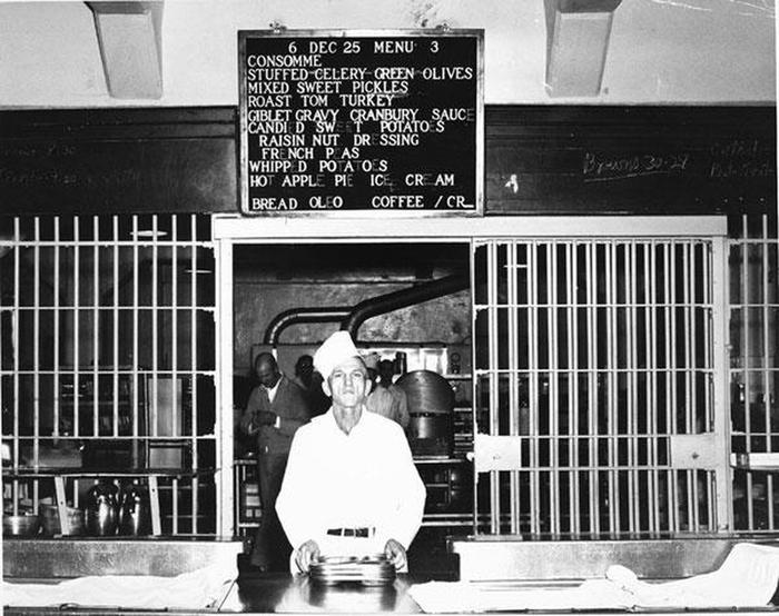 Serving the Christmas Meal, c 1951An inmate worker distributes trays near the steam table. Bars separate the kitchen from the mess hall.  The day’s menu, the Christmas meal, appears on a sign over his head. The menu includes consomme, stuffed celery, green olives and mixed sweet pickles,