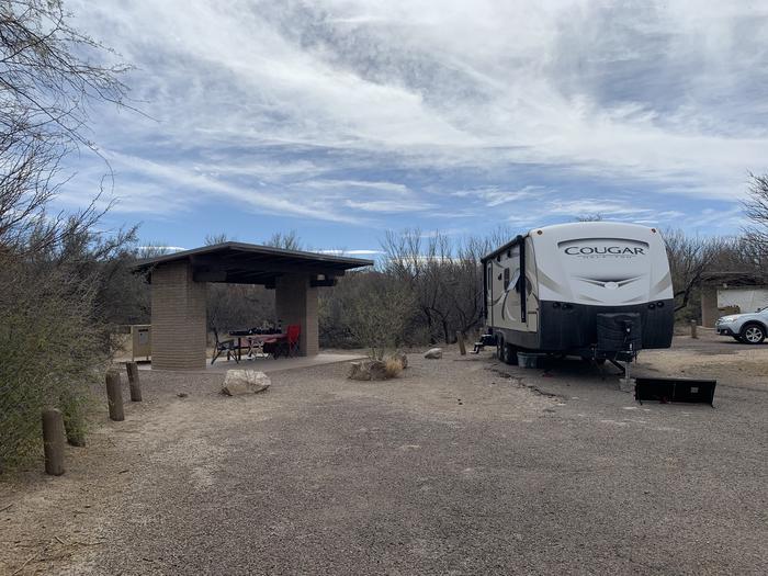A large trailer is parked beside a shade shelter. Slide outs from the trailer are extended out and a solar panel is gathering light from the driveway. There is a large parking space beside the trailer for an additional vehicle to park. View of Site 13 with a parked trailer. 