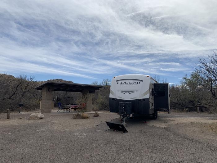 A large trailer is parked beside a shade shelter. Slide outs from the trailer are extended out and a solar panel is gathering light from the driveway. There is still ample room in front of the trailer for additional vehicles to be parked. View of Site 13 with a parked trailer