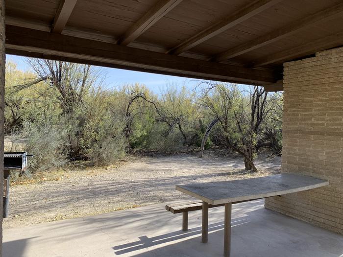 View of the picnic table inside the shade shelter. The table and bench are attached to a wall of the shelter, with one side of the bench open for portable chairs. High bushes and trees surround the shelter, offering additional shade and privacy.View of the shade shelter for Site #16.