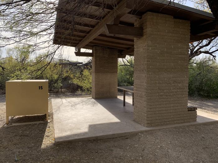 View of the backside of the shade shelter. The shelter is built on to a large, concrete platform and has picnic table built into the walls underneath the roof. A bear box is right beside the concrete platform to allow ease of access to all food and scented items. Bushes and trees grow, surrounding the shelter and providing additional shade and privacy during the day.  Close-up view of the shade shelter.