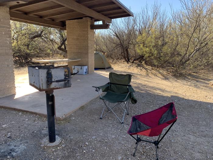 Close-up view of the shade shelter and surrounding area in Site #10. Shown in this picture, campers have setup two portable chairs in front of a metal gril next to the shade shelter. Inside the shelter, there is a picnic table and bench attached to one of the inner walls. Just right behind the shelter is a small, two-person tent. Medium bushes grow around the area to provide additional privacy.Close-up view of the shade shelter area.