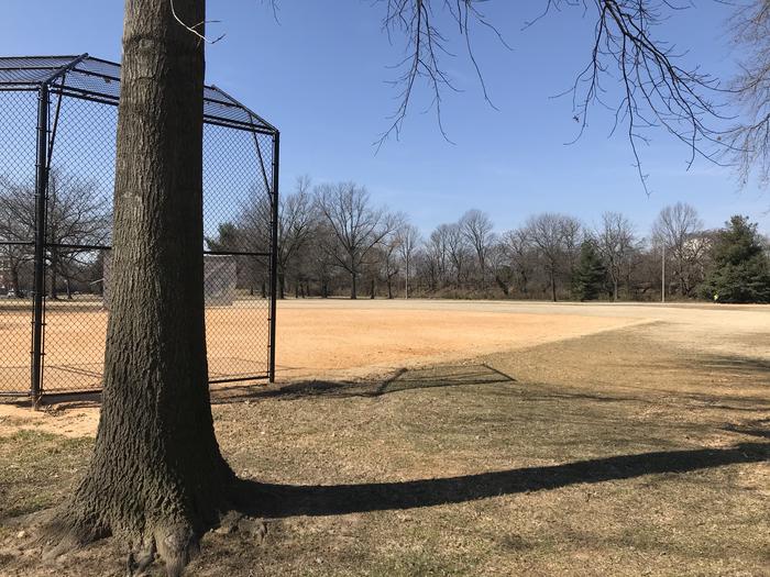 The photo shows field S10 with a tree behind a backstop, infield, and grassy outfield. There are scattered trees in the background.Field S10