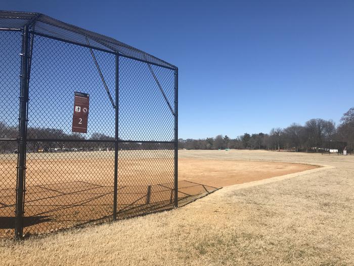 The photo shows a softball field with a backstop, infield, and grassy outfield. There are scattered trees in the background.Field S2