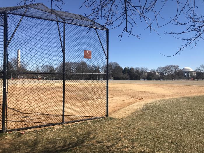 The photo shows a softball field with a backstop, infield, and grassy outfield. The Washington Monument, Thomas Jefferson Memorial, and scattered trees are visible in the background.Field S3