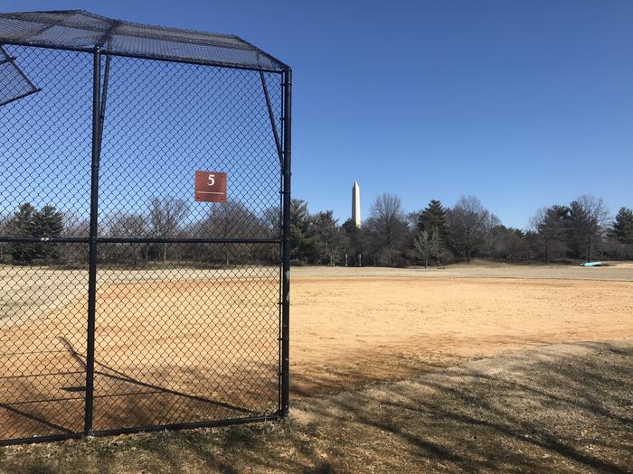 The photo shows a softball field with a backstop, infield, and grassy outfield. The Washington Monument and scattered trees are visible in the background.