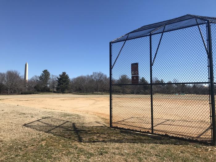 The photo shows a softball field with a backstop, infield, and grassy outfield. The Washington Monument and scattered trees are visible in the background.Field S6