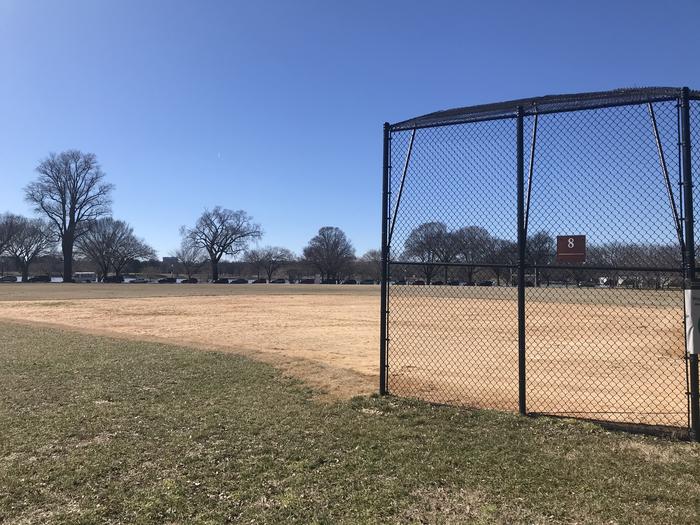 The photo shows a softball field with a backstop, infield, and grassy outfield. Scattered trees are visible in the background.