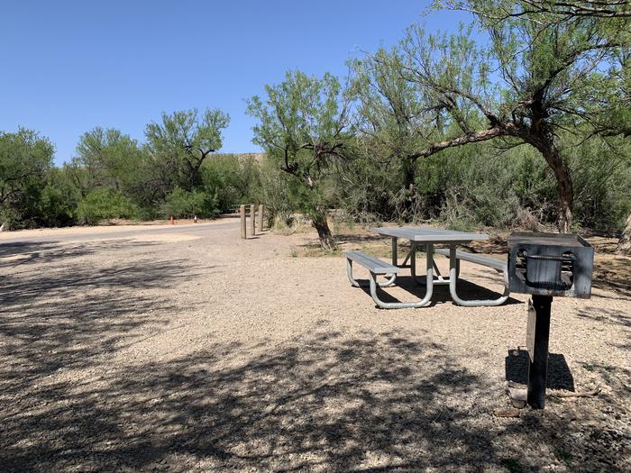 View from the main site area looking out across the pull-through parking space to the main road. There is a picnic table and metal grill in the site, and a long stretch of gravel road for the parking area. Wooden posts at the far end of the driveway mark the angle for the driveway, and protect vehicles from a small irrigation canal that runs adjacent to the site.View of the campsite area and parking space leading to the main road
