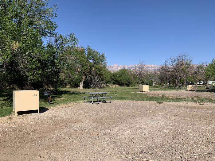 View of the main campsite area. There is a bear box, metal grill, and picnic table on the edge of the site. A large, clear gravel area in the site provides plenty of room for additional parking. A grassy field with trees growing adjacent to the site can be seen, with other neighboring sites and bear boxes in the distance. The Sierra del Carmen mountains glow on the horizon. View of the main campsite area with neighboring campsites