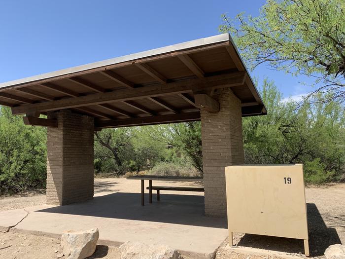 Close-up view of the shade shelter and bear box from the pull-through parking space. The shade shelter is built on a concrete platform and has a picnic table attached underneath the shelter. The bear box isjust outside of the shelter. Large, cleared spaces surround the backside of the shelter, offering plenty of space for pitching tents and shelters. Close-up view of the shade shelter and bear box