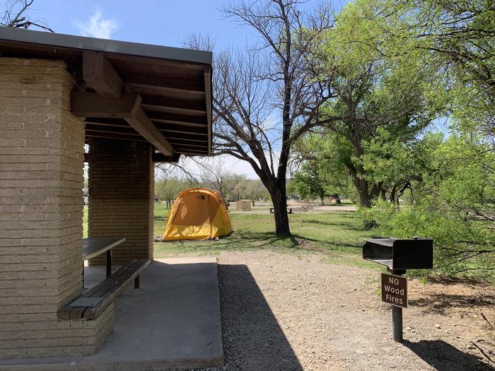 Close-up view of the shade shelter. There is a metal grill adjacent ot the shelter and a large, grassy field behind the site for pitching tents and shelters. A camper has pitched a large, yellow tent in the filed nearby.Close-up view of the shade shelter for Site 20 