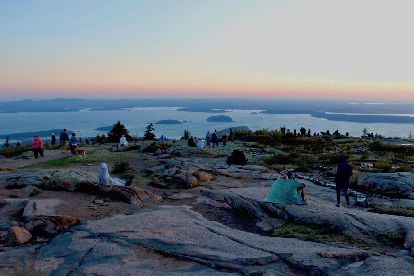 People standing and sitting across granite ledges on top of Cadillac Mountain viewing sunrise with a bay spotted with islands in the background below.Sunrise on Cadillac Mountain.