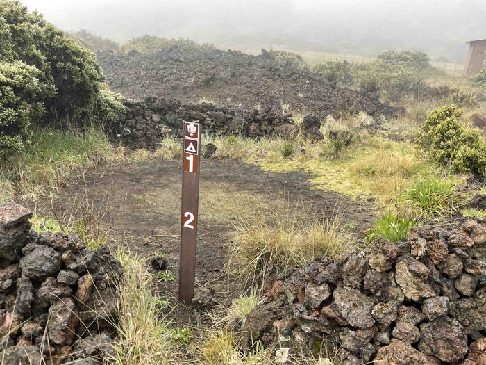 dirt patch surrounded by rocks, grass, shrubs and a brown stake with number 1, 2Hōlua 1 Tent Site without tent