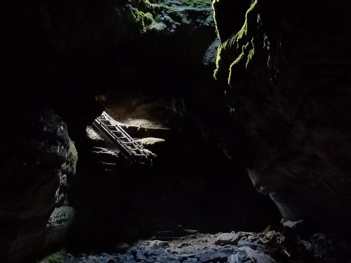 A dark cave has light trickling in from outside, illuminating a small ladder.The exit ladder from the Upper Cave is shown with light from one of the few skylight holes in Ape Cave. 