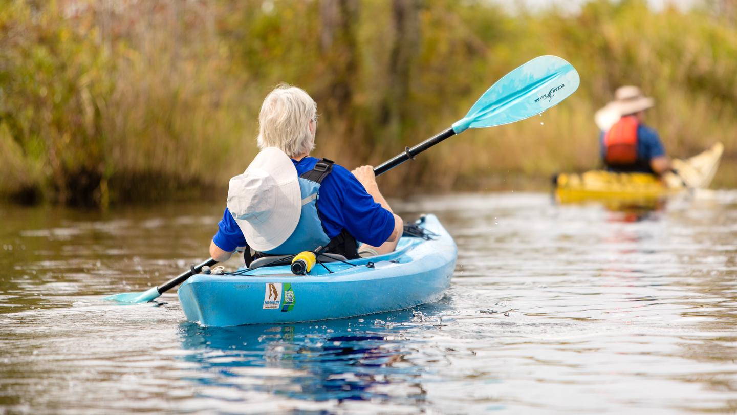 Kayaker in a blue kayak with a black and blue paddle. Second kayaker in background in a yellow kayak, wearing an orange life jacket and a brown hat.Paddling down the Suwannee Canal