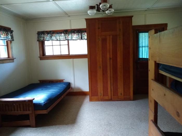A tall cabinet next to a futon bed, with a bunkbed frame in the foreground.Deer Park Cabin's sleeping area.