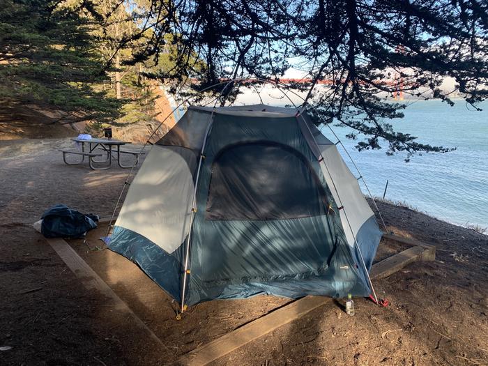 A two person tent is set up in a tent pad near a picnic table with the Golden Gate Bridge in the background.