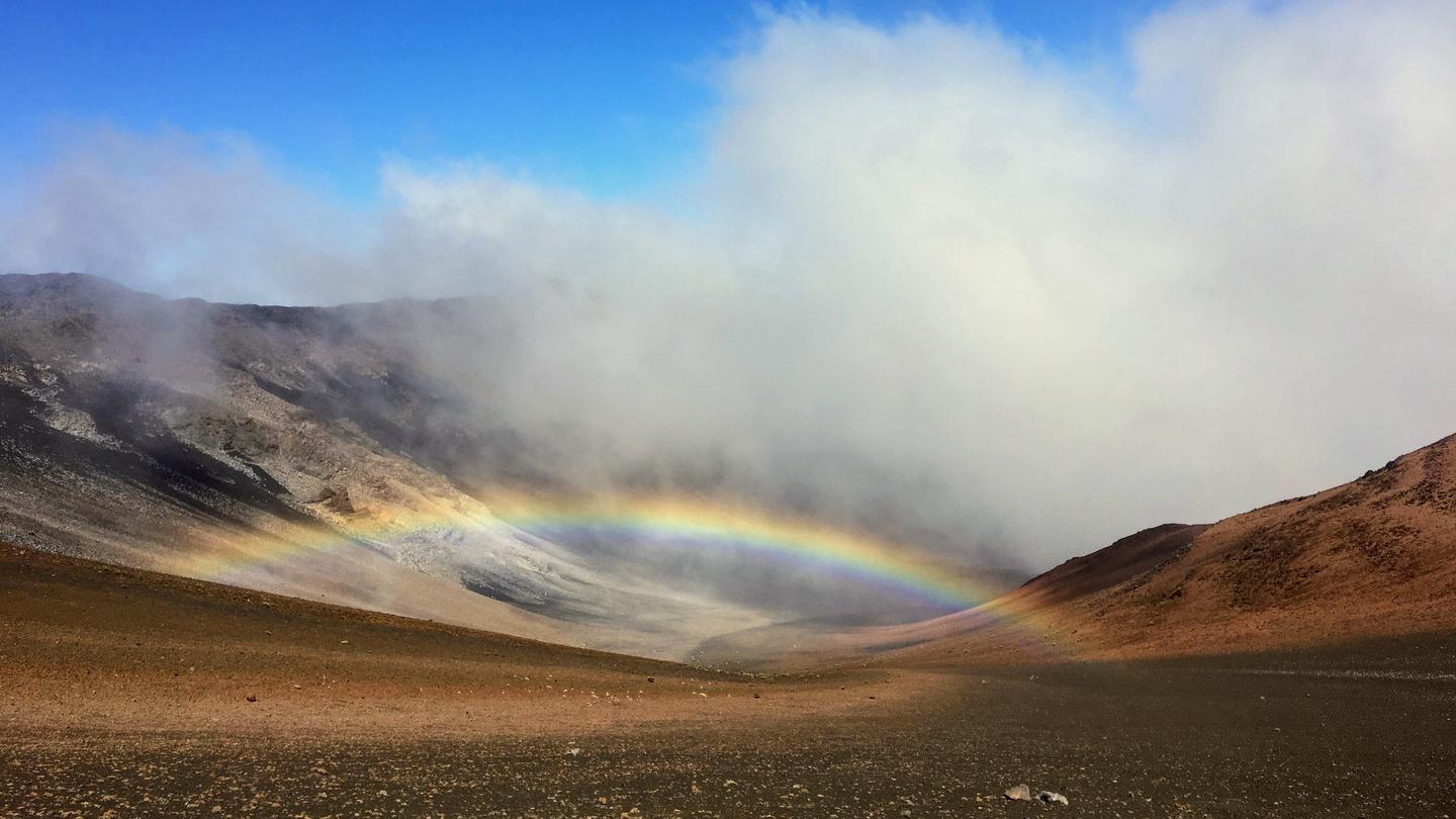 view of crater, orange and black cinder hills with clouds and a low rainbow Wilderness cabins are located in the crater of Haleakalā National Park.