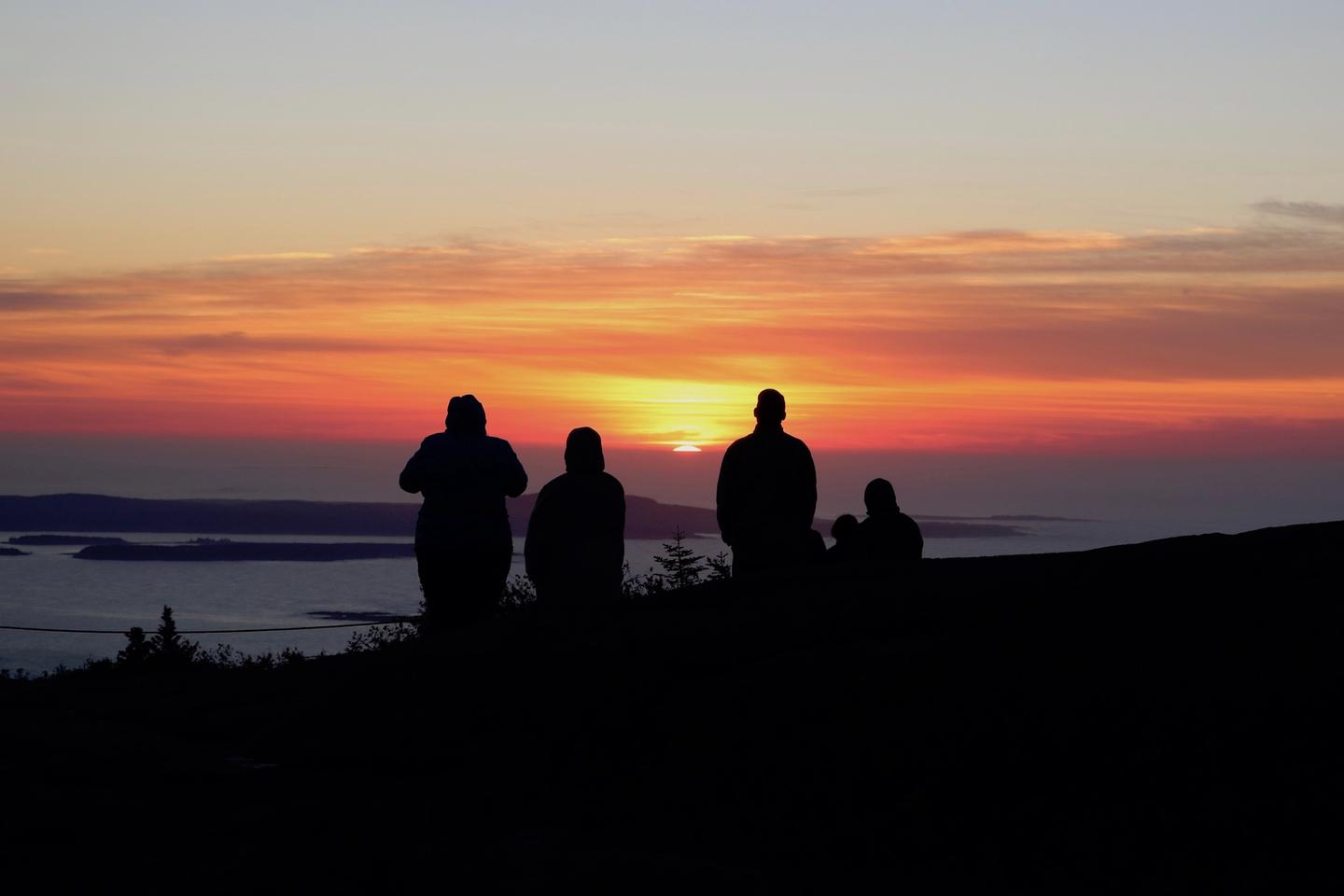 Five people shown in silhouette watch the bright orange and red sunrise on the horizon overlooking the ocean from a mountain top.Sunrise on Cadillac Mountain.