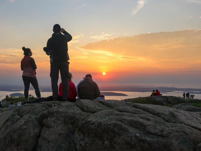About a dozen people sitting and standing across granite outcrops watch the bright orange and red sunrise on the horizon overlooking the ocean from a mountain top.Sunrise on Cadillac Mountain.
