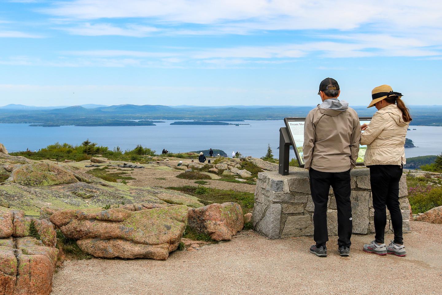 A man and woman dressed in light coats and dark pants with hats stand next to each other in front of an interpretive panel overlooking the ocean and distant mountains from the top of a mountain.Interpretive display at the Cadillac Summit.