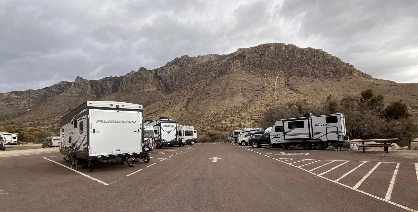 :  A view of RV campground parking area with sites 21-30 shown.  Site 30 is located along the left side of travel lane.  Mountain views in the background.A view of RV campground parking area with sites 21-30 shown. Campground is shown when near capacity. 