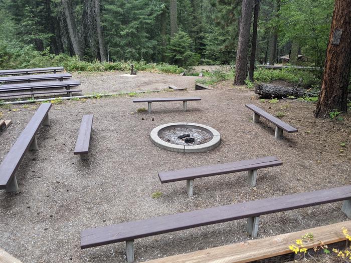 Fire pit surrounded by benches, with picnic tables in the background.Spring Creek Group site fire pit area, and picnic tables.