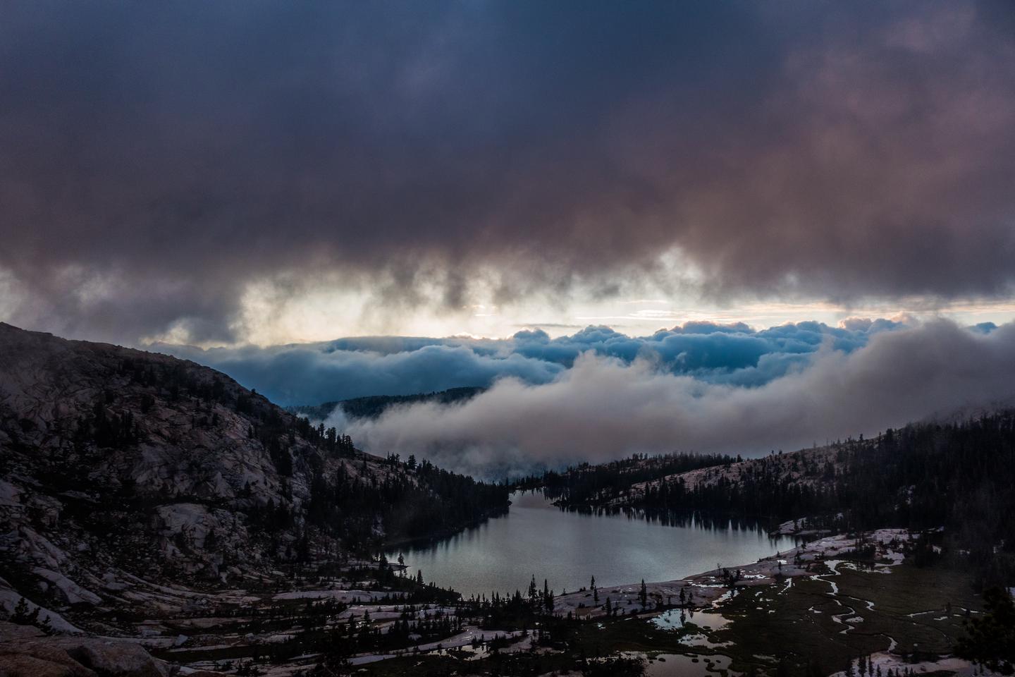 View of a lake below in, with clouds above and clouds below the lake in the distanceA glacial lake sits high in the Sierra, nestled in the heart of a breaking storm. 
