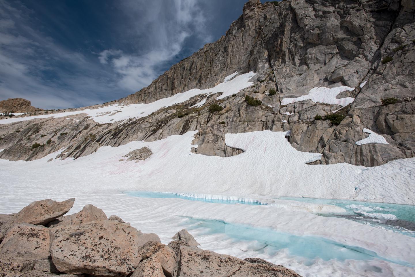 A small ice covered lake at the base of a mountain.A lake high in the Sierra, holding onto ice and snow from the previous winter. In some years, snow can persist in the High Sierra through July. 